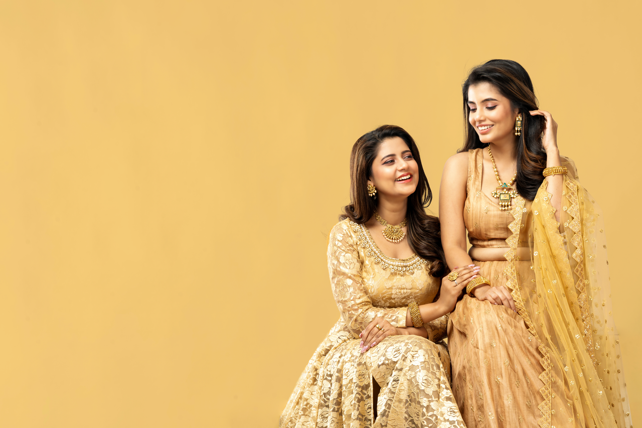 Services Two smiling women in elegant traditional Indian dresses with a warm backdrop.